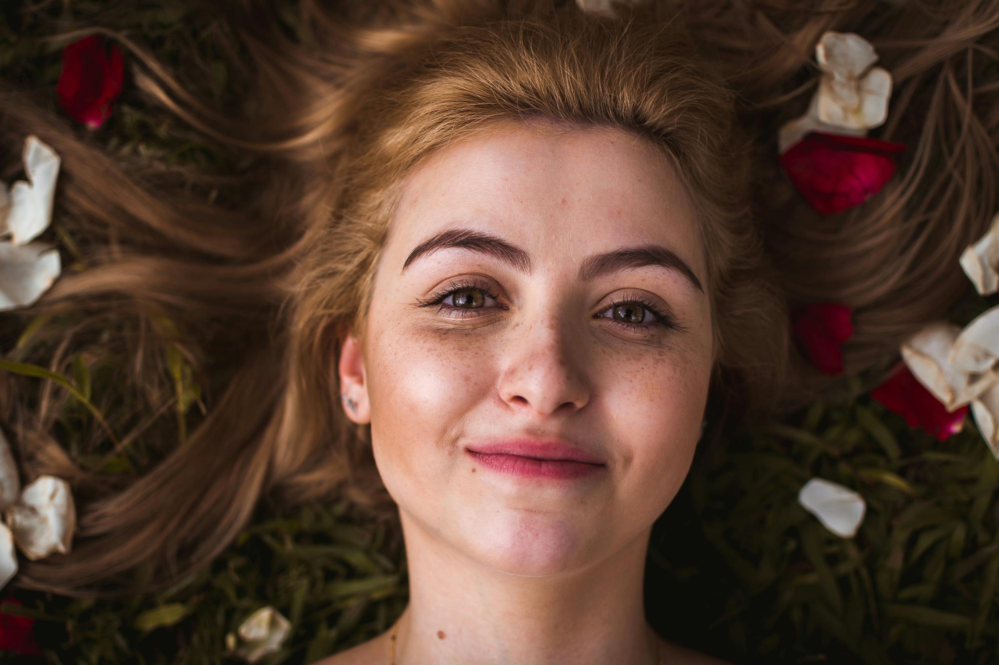 Portrait of a smiling young woman with rose petals in her hair, lying on grass outdoors.